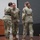 USARIEM COC/Relinquishment of Responsibility 2026 Three U.S. Army soldiers in OCP (Operational Camouflage Pattern) uniforms are gathered on a stage during what appears to be a military ceremony. Two soldiers appear to be embracing or shaking hands while a third stands nearby observing. Military flags and a podium are visible in the background, suggesting a formal change of command or award ceremony.