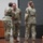 USARIEM COC/Relinquishment of Responsibility 2026 Three U.S. Army soldiers in Operational Camouflage Pattern (OCP) uniforms are gathered on a stage during what appears to be a military ceremony. Two soldiers appear to be engaged in a handshake or embrace while a third observes. Military flags and a unit guidon are visible in the background alongside a wooden podium.
