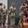 USARIEM COC/Relinquishment of Responsibility 2026 A U.S. Army military ceremony is taking place on a wooden stage, likely a change of command or unit activation/deactivation ceremony. Four soldiers in OCP (Operational Camouflage Pattern) uniforms are present, with one soldier handling a maroon/burgundy unit guidon or colors on a stand. A wooden podium with a military insignia is visible in the background.