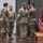 USARIEM COC/Relinquishment of Responsibility 2026 Four U.S. Army soldiers in OCP (Operational Camouflage Pattern) uniforms are gathered on a stage during what appears to be a military ceremony, likely a change of command or award presentation. A podium with a microphone is visible to the left, and multiple military unit flags including the U.S. flag and a maroon guidon are displayed on the right. Two soldiers face each other in conversation while two others stand nearby with their backs to the camera.