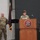 USARIEM COC/Relinquishment of Responsibility 2026 Two U.S. Army soldiers in OCP (Operational Camouflage Pattern) uniforms are participating in an official military ceremony or briefing at USARIEM (U.S. Army Research Institute of Environmental Medicine). One soldier stands at a wooden podium bearing the USARIEM seal and speaks into a microphone, while a second soldier stands at attention nearby. A maroon/dark red military guidon flag is displayed in the background.
