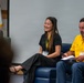 Three individuals are seated at what appears to be a U.S. Army panel or event, with two prominently visible in the foreground. A smiling woman in black clothing and a man in a yellow polo shirt both wear U.S. Army-branded lanyards and name badges. A third man in a gray blazer is partially visible in the background.