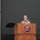 USARIEM COC/Relinquishment of Responsibility 2026 A U.S. Army soldier is speaking at a podium bearing the USARIEM (U.S. Army Research Institute of Environmental Medicine) seal during what appears to be an official military ceremony or briefing. A second soldier stands at attention to the left near a maroon and gold unit guidon or flag. The setting is an auditorium or formal presentation hall.