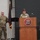 USARIEM COC/Relinquishment of Responsibility 2026 Two U.S. Army soldiers in OCP (Operational Camouflage Pattern) uniforms are participating in an official military ceremony or briefing at a USARIEM (U.S. Army Research Institute of Environmental Medicine) podium. A female soldier is speaking at the wooden lectern bearing the USARIEM logo, while a male soldier stands at attention to her left near a maroon and gold guidon flag. The setting appears to be a formal auditorium or conference room.