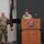 USARIEM COC/Relinquishment of Responsibility 2026 Two U.S. Army soldiers in OCP (Operational Camouflage Pattern) uniforms are participating in an official military ceremony or briefing at a USARIEM (U.S. Army Research Institute of Environmental Medicine) podium. A female soldier is speaking at the wooden lectern while a male soldier stands at attention nearby. A dark maroon/burgundy military guidon flag is displayed in the background.