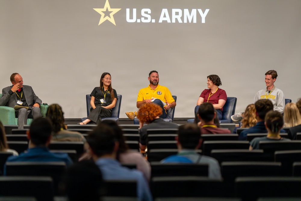 A panel discussion is taking place on a stage beneath a large U.S. Army logo and star emblem. Five panelists are seated in blue chairs on an elevated stage, facing an audience seated in tiered auditorium-style seating. The event appears to be a U.S. Army-sponsored forum or recruitment/informational event.