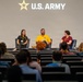 A panel discussion is taking place on a stage beneath a large U.S. Army logo and star emblem. Five panelists are seated in blue chairs on an elevated stage, facing an audience seated in tiered auditorium-style seating. The event appears to be a U.S. Army-sponsored forum or recruitment/informational event.
