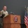 USARIEM COC/Relinquishment of Responsibility 2026 A U.S. Army soldier in camouflage uniform is speaking at a podium bearing the USARIEM (U.S. Army Research Institute of Environmental Medicine) seal. The setting appears to be a formal military ceremony or briefing, with multiple flags displayed to the right of the podium. The speaker is holding documents and addressing an audience from the wooden lectern.