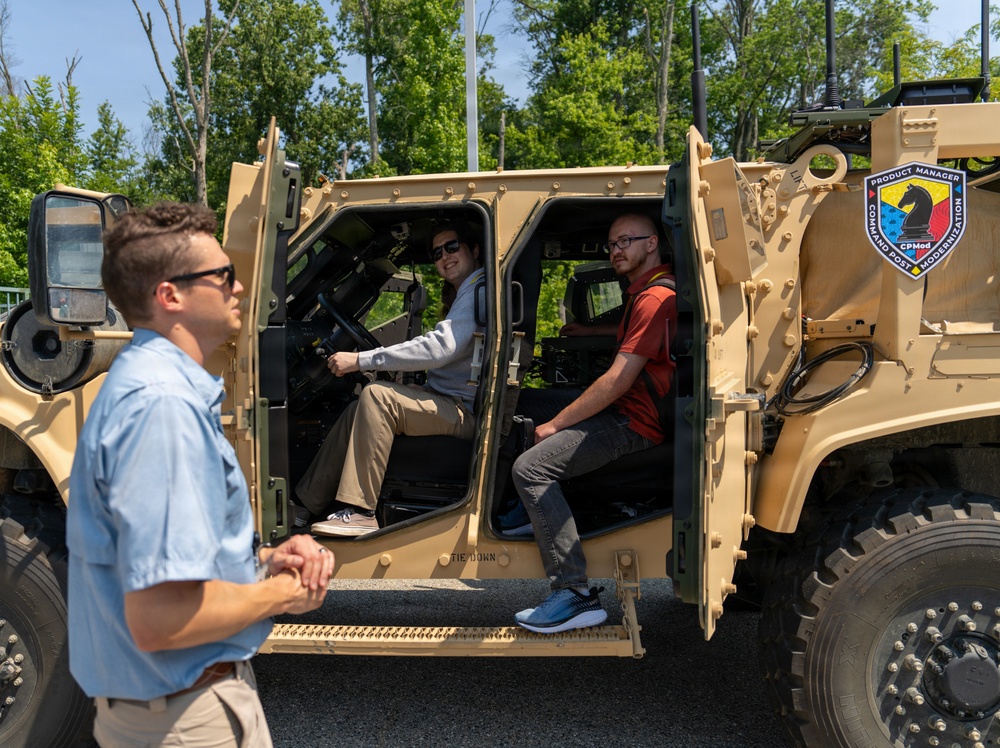 Three civilians are examining a tan military tactical vehicle bearing a 'Product Manager Command Post Modernization' (CPMod) insignia. Two individuals are seated inside the open-doored vehicle while a third stands outside observing. The setting appears to be an outdoor demonstration or equipment showcase event on a paved surface.