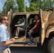 Three civilians are examining a tan military tactical vehicle bearing a 'Product Manager Command Post Modernization' (CPMod) insignia. Two individuals are seated inside the open-doored vehicle while a third stands outside observing. The setting appears to be an outdoor demonstration or equipment showcase event on a paved surface.
