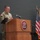 USARIEM COC/Relinquishment of Responsibility 2026 A U.S. Army soldier in camouflage uniform is speaking at a podium bearing the USARIEM (U.S. Army Research Institute of Environmental Medicine) seal. The setting appears to be an official military ceremony or briefing, with multiple flags displayed to the right of the podium. The soldier appears to be addressing an audience from the elevated stage.