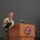 USARIEM COC/Relinquishment of Responsibility 2026 A U.S. Army soldier in camouflage uniform is speaking at a podium bearing the USARIEM (U.S. Army Research Institute of Environmental Medicine) seal. The soldier appears to be delivering a speech or remarks at an official military ceremony or event. Several flags, including the U.S. flag and military unit colors, are displayed in the background.