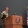 USARIEM COC/Relinquishment of Responsibility 2026 A U.S. Army officer in camouflage uniform is speaking at a podium bearing the USARIEM (U.S. Army Research Institute of Environmental Medicine) seal during an official military ceremony or event. Multiple flags, including the U.S. flag and military unit colors, are displayed to the right of the podium. The setting appears to be an indoor auditorium or conference hall.