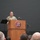 USARIEM COC/Relinquishment of Responsibility 2026 A U.S. Army soldier in OCP (Operational Camouflage Pattern) uniform is speaking at a podium bearing the USARIEM (U.S. Army Research Institute of Environmental Medicine) logo during an official military ceremony or event. An audience of military personnel is seated in the foreground, while another soldier stands at attention near a unit guidon flag on the left. An American flag is partially visible on the right side of the frame.