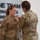 USARIEM COC/Relinquishment of Responsibility 2026 A military pinning ceremony is taking place in what appears to be an official building or headquarters. One soldier, identified by the name tape 'ROSSER,' is receiving a pin or insignia on their uniform from another soldier. A third uniformed individual stands nearby holding what appears to be a document or certificate.