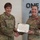 USARIEM COC/Relinquishment of Responsibility 2026 Two U.S. Army female soldiers are posing together while jointly holding a certificate during what appears to be a formal award ceremony. The soldier on the right is wearing a Legion of Merit medal on her uniform, indicating she is the award recipient. The background displays a partial motivational phrase reading 'ONE TEAM... ADVANCING... INNOVATIVE... LOGY... DAY & RROW...'