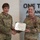 USARIEM COC/Relinquishment of Responsibility 2026 Two U.S. Army female soldiers are posing together while jointly holding a certificate during what appears to be a formal award presentation ceremony. The soldier on the right is wearing a Legion of Merit medal (red and white ribbon with gold star) on her uniform. The setting appears to be an official military facility with a motivational slogan visible on the wall behind them.