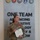 A U.S. Army Colonel (identified by eagle rank insignia on collar) named Rosser stands before a joint military organizational display wall reading 'ONE TEAM ADVANCING INNOVATIVE TECHNOLOGY &amp; KNOW[HOW]'. She is holding a military certificate and wearing a medal on her uniform, suggesting this is an awards or recognition ceremony. The background features seals of multiple U.S. military branches.