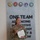 A U.S. Army soldier, identified by name tape as 'ROSSER,' stands in front of a military display board reading 'ONE TEAM ADVANCING INNOVATIVE TECHNOLOGY &amp; KNOW[LEDGE].' She is holding a certificate or award document and wearing a medal on her uniform, suggesting this is an award or recognition ceremony. Five military branch seals are visible above the text on the display board.