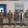 USARIEM COC/Relinquishment of Responsibility 2026 Seven individuals are posed in a formal group photo inside what appears to be a military facility. Three are wearing U.S. Army combat uniforms (ACU/OCP pattern), while four are in civilian attire. They are standing in front of a wall-mounted plaque reading 'ONE TEAM' with military branch seals, alongside a large display screen.