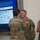 USARIEM COC/Relinquishment of Responsibility 2026 A military award ceremony is taking place indoors, where a uniformed soldier is receiving a medal or decoration being pinned to their chest by another uniformed individual. A civilian woman in a floral dress observes the ceremony alongside another soldier holding a folder or certificate. The event appears to be occurring in front of a DEVCOM presentation screen displaying a 'Partnerships' slide.