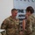 USARIEM COC/Relinquishment of Responsibility 2026 A U.S. Army military awards ceremony is taking place indoors, likely at a command or administrative facility. A soldier identified by name tape as 'MARCONI' is receiving a medal from a female officer, while another soldier holds an award certificate. The background signage partially reads 'ONE TEAM' with additional text obscured.