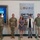 USARIEM COC/Relinquishment of Responsibility 2026 Seven individuals are standing in a line inside what appears to be a U.S. military facility or research center. Three individuals are wearing U.S. Army combat uniforms (OCP/multicam), while four civilians stand alongside them. A prominent wall plaque reading 'ONE TEAM' with military branch seals is visible in the background, along with a large display screen.