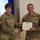 USARIEM COC/Relinquishment of Responsibility 2026 Two U.S. Army soldiers in OCP (Operational Camouflage Pattern) uniforms are shaking hands during a military award ceremony. The male soldier, identified by his name tape as 'MARCONI,' is receiving a Legion of Merit certificate and is wearing the corresponding medal on his chest. A civilian woman is partially visible on the right edge of the frame.