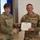 USARIEM COC/Relinquishment of Responsibility 2026 Two U.S. Army soldiers are shaking hands while jointly holding a certificate from the United States of America, indicating an award or commendation ceremony. The male soldier on the right, identified by his name tape as 'MARCONI' with the rank of Staff Sergeant, is wearing a red medal on his chest. A civilian woman is partially visible on the right edge of the frame.