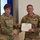 USARIEM COC/Relinquishment of Responsibility 2026 Two U.S. Army soldiers are shaking hands during a formal award ceremony. The soldier on the right, identified by nametape as 'MARCONI,' is receiving a certificate and is wearing a Legion of Merit medal on his chest. A civilian woman is partially visible on the right edge of the frame.