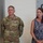 USARIEM COC/Relinquishment of Responsibility 2026 A U.S. Army soldier wearing OCP (Operational Camouflage Pattern) uniform with the name tape 'MARCONI' and a medal pinned to his chest stands beside a civilian woman in a blue lace dress. The setting appears to be an indoor military or government facility, likely during an awards or recognition ceremony. A partial wall display with text reading 'ONE T... ADVAN... INNOV... TECHN... R TO... OMOR...' is visible in the background.