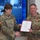USARIEM COC/Relinquishment of Responsibility 2026 Two U.S. Army soldiers are shaking hands while jointly holding a certificate in a formal military recognition ceremony. The soldier on the right is wearing a medal (appears to be a Meritorious Service Medal or similar decoration) pinned to his uniform. The ceremony is taking place indoors in front of a large display screen showing a digital/simulation environment.