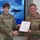 USARIEM COC/Relinquishment of Responsibility 2026 Two U.S. Army soldiers in MultiCam/OCP camouflage uniforms are shaking hands while jointly holding a certificate in a red folder. The soldier on the right is wearing a medal on their chest, suggesting this is an award or recognition ceremony. The setting appears to be an indoor military facility with a large digital display screen in the background.