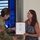 USARIEM COC/Relinquishment of Responsibility 2026 A U.S. Army soldier in camouflage uniform is presenting a formal certificate or award to a civilian woman during what appears to be an official military ceremony. The certificate appears to be an honorary membership document from the United States Army Medical Department Regiment. The event is taking place indoors in front of a large display screen showing military doctrine text.