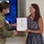 USARIEM COC/Relinquishment of Responsibility 2026 A U.S. Army soldier in OCP (Operational Camouflage Pattern) uniform is presenting a framed certificate to a civilian woman during a formal recognition ceremony. The certificate appears to be an Honorary Member designation from the United States Army Medical Department Regiment. The event is taking place indoors in front of a large display screen showing military briefing content.