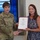 USARIEM COC/Relinquishment of Responsibility 2026 A U.S. Army officer, identified by name tape as 'JENKINS' and wearing a one-star General rank insignia, presents a certificate to a civilian woman in a blue lace dress. The two are shaking hands while jointly holding the framed certificate in what appears to be a military briefing room. The certificate appears to be an honorary membership or recognition document.