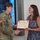 USARIEM COC/Relinquishment of Responsibility 2026 A U.S. Army soldier in OCP (Operational Camouflage Pattern) uniform is presenting a Certificate of Appreciation to a civilian woman in a blue floral dress. The two individuals are shaking hands while jointly holding the certificate during what appears to be a formal recognition ceremony. The event takes place indoors in front of a large display screen.