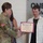 USARIEM COC/Relinquishment of Responsibility 2026 A U.S. Army officer in camouflage uniform is presenting a certificate to a young civilian man during what appears to be a formal recognition ceremony. The certificate is visibly labeled 'B·R·A·T' from the United States Army. Several other individuals, including another uniformed soldier identified by a 'MARCONI' name tape, are present in the background.