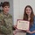 USARIEM COC/Relinquishment of Responsibility 2026 A U.S. Army officer in camouflage uniform is presenting a certificate to a young woman during what appears to be a formal recognition ceremony. The certificate is from 'The United States Army B.R.A.T.' program, awarded to 'Adelyn E. Marconi.' A young man in the background holds a similar certificate, suggesting multiple recipients are being recognized.