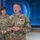 USARIEM COC/Relinquishment of Responsibility 2026 A U.S. Army Sergeant Major named Marconi stands at the center of the image wearing an OCP (Operational Camouflage Pattern) uniform and a red medal, likely the Meritorious Service Medal or similar decoration, suggesting a military awards ceremony. Behind him stands a female U.S. Army soldier, and to the sides are partially visible civilians and military personnel. The setting appears to be an indoor military facility with a large digital display screen in the background.