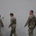 USARIEM COC/Relinquishment of Responsibility 2026 Three military personnel in matching camouflage uniforms (OCP/MultiCam pattern) are walking in a line through what appears to be an indoor facility. Two appear to be female and one male, all wearing the same pattern of Army Combat Uniform. A civilian individual is partially visible on the left edge of the frame.