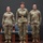 USARIEM COC/Relinquishment of Responsibility 2026 Three U.S. Army soldiers in Operational Camouflage Pattern (OCP) uniforms stand at attention on a stage or platform, likely during a formal military ceremony. The center soldier is distinguished by a dress belt with a gold buckle, suggesting a position of higher rank or a ceremonial role. Two blue chairs are visible in the background behind them.