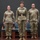 USARIEM COC/Relinquishment of Responsibility 2026 Three U.S. Army soldiers in Operational Camouflage Pattern (OCP) uniforms stand at attention on a raised stage or platform. The center figure appears to be of higher rank, distinguished by a belt with a prominent buckle. This appears to be a formal military ceremony or official event.