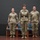 USARIEM COC/Relinquishment of Responsibility 2026 Three U.S. Army soldiers in Operational Camouflage Pattern (OCP) uniforms stand at attention on a stage during what appears to be a formal military ceremony. The center figure is distinguished by a black belt with a brass buckle, suggesting a position of higher rank or ceremony role. A maroon flag with a medical caduceus emblem is visible on the left, indicating a U.S. Army Medical Command affiliation.