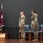 USARIEM COC/Relinquishment of Responsibility 2026 Three U.S. Army soldiers in OCP (Operational Camouflage Pattern) uniforms stand at attention on a stage during what appears to be a formal military ceremony. A display of flags is visible to their left, including the U.S. national flag and maroon unit guidons with military insignia. Three chairs are positioned behind the soldiers on the stage.