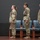 USARIEM COC/Relinquishment of Responsibility 2026 Three U.S. Army soldiers in Operational Camouflage Pattern (OCP) uniforms stand at attention on a stage during what appears to be a formal military ceremony. Two female soldiers and one male soldier are positioned in a line facing left, with empty ceremonial chairs behind them. A maroon military guidon or unit flag is visible on the left side of the frame.