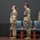 USARIEM COC/Relinquishment of Responsibility 2026 Three U.S. Army soldiers in OCP (Operational Camouflage Pattern) uniforms stand at attention on a stage or platform during what appears to be a formal military ceremony. Two female soldiers and one male soldier are positioned in a line facing the same direction, with blue chairs and unit guidon flags visible in the background. The maroon/burgundy flags suggest an affiliation with the U.S. Army Medical Command or a medical unit.