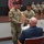 USARIEM COC/Relinquishment of Responsibility 2026 Two U.S. Army soldiers in OCP (Operational Camouflage Pattern) uniforms are presenting a gift bag to a seated civilian man during what appears to be a military ceremony or retirement event. The setting is an auditorium or assembly hall with flags and additional uniformed personnel seated on a stage in the background. A civilian woman is also seated nearby, suggesting this may be a presentation to a family member or retiring service member.