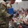 USARIEM COC/Relinquishment of Responsibility 2026 Two U.S. Army soldiers in MultiCam/OCP camouflage uniforms are presenting gifts, including flowers and a blue gift bag, to seated civilians in what appears to be an auditorium or meeting room. The audience consists of approximately 15-20 people of varying ages seated in teal/green auditorium chairs. The setting suggests a formal military ceremony, possibly a promotion, retirement, or unit recognition event.