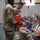 USARIEM COC/Relinquishment of Responsibility 2026 Two U.S. Army soldiers in OCP (Operational Camouflage Pattern) uniforms are presenting gift bags to seated civilians in what appears to be an auditorium or ceremony hall. The event appears to be a formal military ceremony, likely a promotion, retirement, or recognition event, as evidenced by the dressed audience and flowers held by one attendee. State or organizational flags are visible hanging in the background.
